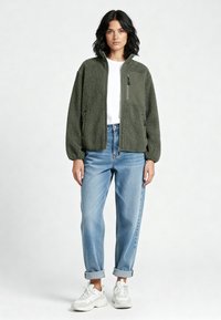 Donna in piedi che indossa una giacca in pile verde oliva con cerniera, una camicia bianca, jeans azzurri arrotolati e scarpe da ginnastica bianche su uno sfondo neutro.