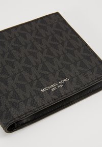Portefeuille noir en matériau texturé avec un motif géométrique. L'avant affiche un logo métallique "MICHAEL KORS" avec "EST. 1981."