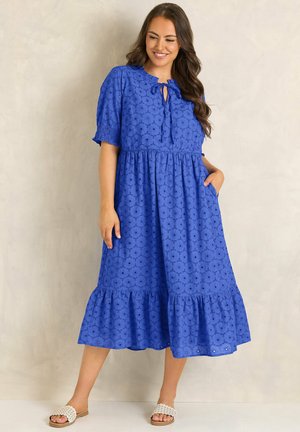 Uśmiechnięta kobieta stoi w niebieskiej sukience midi z ażurowym wzorem, krótkimi rękawami, falbanianym dołem i białymi klapkami.