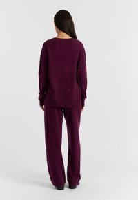 Lila gestrickter Pullover und passende weit geschnittene Hosen, die eine entspannte Passform und weiche Textur bieten, von hinten betrachtet. Glattes Gewebe, minimale Details.