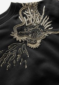 Sudadera negra con intrincado bordado metálico de cuentas y lentejuelas con un diseño de dragón, que presenta texturas variadas y elementos reflectantes.