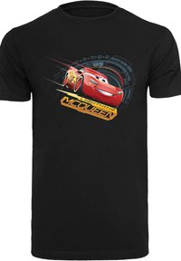 Melnā T-kreklā ir attēlots grafikas attēls ar Lightning McQueen sarkanu krāsu un ziliem acīm, ko ieskauj ātruma līnijas un digitāla dizaina elementi.