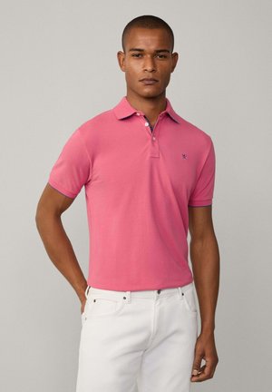 Lyserød polo skjorte med krave; korte ærmer; kontrasterende navy detaljer på kraven. Modellen bærer hvide jeans. Glat bomuldsstruktur.
