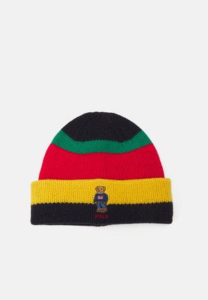 Gebreide muts met zwarte, groene, rode en gele strepen, met een kleine geborduurde beer die een trui en spijkerbroek draagt met daaronder de tekst "POLO".