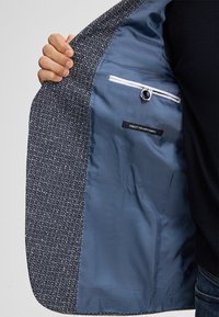 Marineblau strukturierte Jacke mit einer glatten blauen Fütterung, ausgestattet mit einer Innentasche und einem Etikett, das "FEINSTES ITALIENISCHES STOFF" liest.