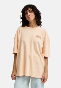 Απαλό ροδακινί βαμβακερό T-shirt με κοντά μανίκια, με στρογγυλή λαιμόκοψη και κεντητή λεπτομέρεια στο στήθος. Χαλαρή εφαρμογή με απαλό υφή.