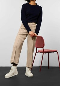 Marinblå tröja, beige byxor och vita högskurna sneakers. Byxorna har framfållar. Bakgrunden inkluderar en rosa stol på ett svart golv.