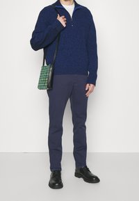 Marineblauer Strick-Zipper-Pullover mit blauer Kragen, kombiniert mit marineblauen Hosen und schwarzen Schuhen. Eine kleine grün-karierte Umhängetasche rundet das Outfit ab.