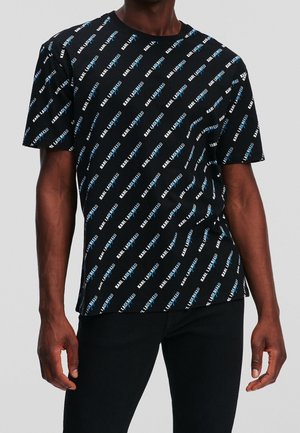 Czarny t-shirt z krótkim rękawem z powtarzającym się ukośnym białym i niebieskim napisem "KARL LAGERFELD", noszony przez osobę o ciemnej karnacji w czarnych spodniach.