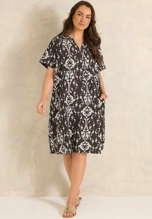 Kobieta ubrana w czarno-białą, wzorzystą sukienkę do kolan z krótkimi rękawami i sandały, stojąca na neutralnym tle.