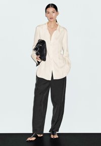 Camicia button-up crema, pantaloni neri larghi e sandali neri flat. Modella che tiene una borsa nera con dettagli in argento. Design semplice e sartoriale.