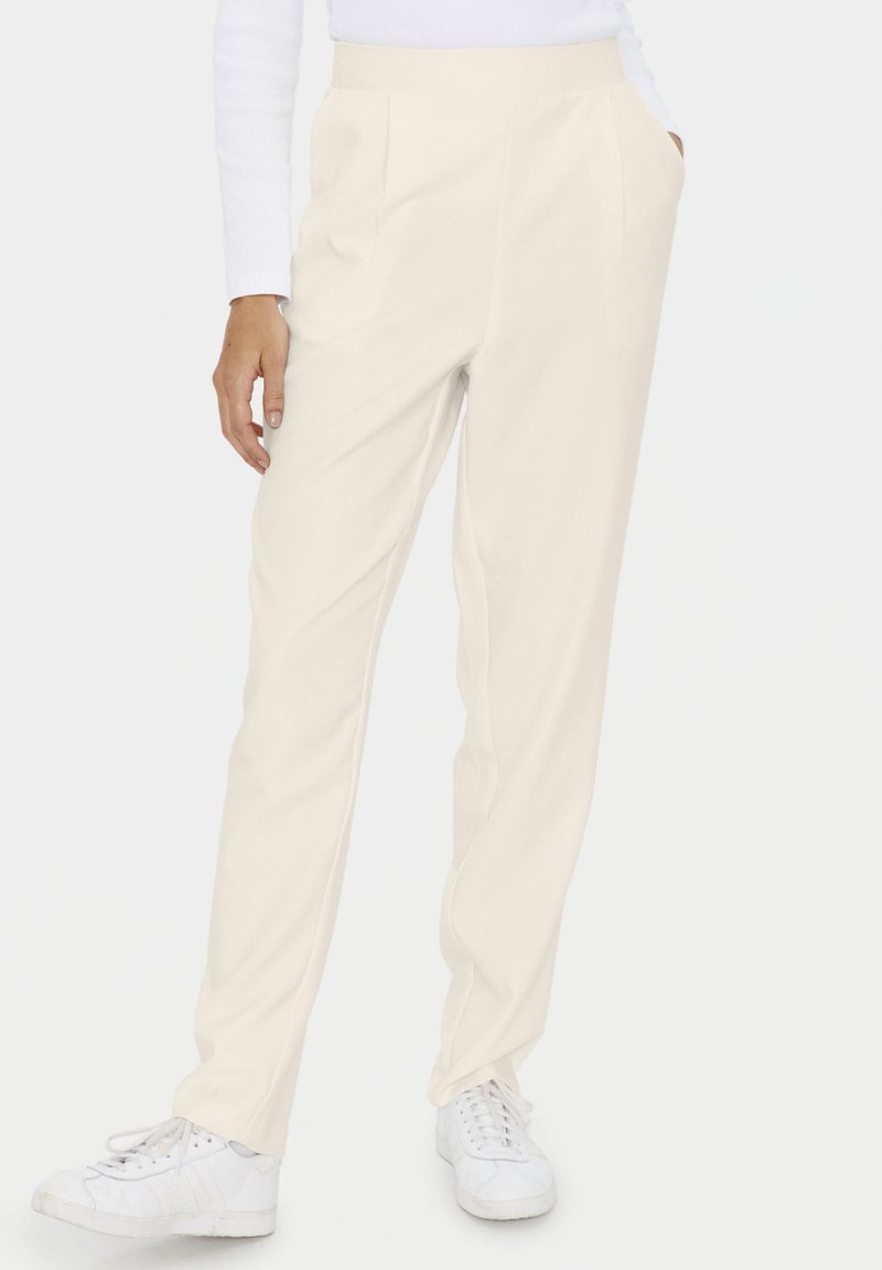 Femme portant un pantalon crème taille haute avec poches latérales et des baskets blanches sur un fond uni.