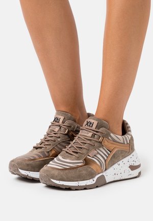 Jalat, joissa on ruskeat ja beiget Xti-tennarit, joissa on seepraraidat, nupukkinahkaa ja valkoiset pilkulliset pohjat valkoisella taustalla.