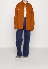 Chemise-veston marron surdimensionnée, t-shirt blanc en dessous, jean cargo bleu marine taille haute, et chaussures slips-on à motifs.