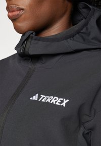 Svart Terrex-jacka med dragkedja framtill, texturerad tyg och en vit broderad logotyp på bröstet. Har en åtsittande huva och krage.