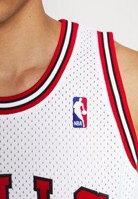 Jerseu de baschet alb cu textură din plasă, având un guler ribonat cu dungi roșii și negre și un patch cu logo-ul NBA albastru pe piept.