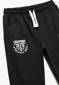 Svarta sweatpants med elastisk midja, med vita dragsnören och en vit grafisk trycketikett på vänster lår.