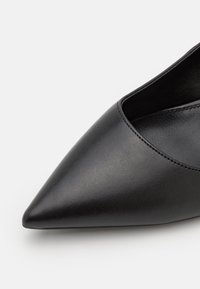 Chaussure noire en cuir à bout pointu avec un design élégant, présentant un détail découpé sur le côté et une texture lisse. Couture minimale visible.