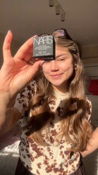 Young woman with long hair holds NARS Soft Matte concealer close to the camera, smiling in warm natural sunlight indoors.