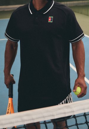 Personne portant une tenue de tennis noire tenant une balle de tennis jaune et une raquette de tennis orange sur un court de tennis bleu derrière le filet.