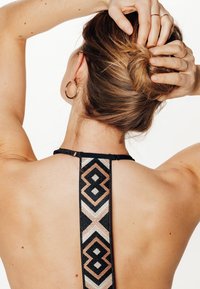 Haut halter noir avec des bretelles décoratives à motif géométrique en noir, blanc et rose doré métallique. Cheveux coiffés en chignon, montrant des boucles d'oreilles.