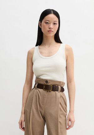 Kobieta o długich czarnych włosach, ubrana w biały, bezrękawnik, beżowe spodnie z wysokim stanem oraz brązowy pasek ze złotą klamrą.