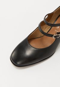 Scarpa in pelle nera con punta squadrata, caratterizzata da due cinturini incrociati e una fibbia in argento. L'interno è color cammello, con una texture liscia.