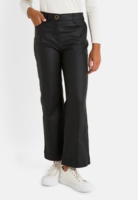 Mujer vistiendo pantalones anchos de cintura alta negros con botones y bolsillos, combinados con zapatillas blancas y una blusa de manga larga color crema.