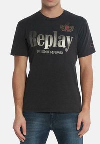 Camiseta negra de cuello redondo hecha de algodón con texto metálico plateado "Replay" y un acento de escudo rojo. Mangas cortas y un corte relajado.