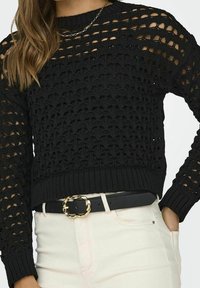Schwarzer Strickpullover mit strukturiertem, offenem Gewebe-Design und langen Ärmeln. Kombiniert mit einem schwarzen Gürtel und hellen Hosen.
