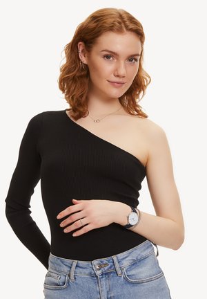 Top nero a coste con spalla scoperta e manica lunga su un lato, abbinato a jeans azzurri a vita alta, con orologio d'argento e anelli.