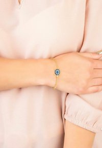 Bracciale in oro con una pietra blu centrale, circondata da piccoli dettagli blu, indossato su un polso scoperto su un morbido tessuto di colore chiaro.