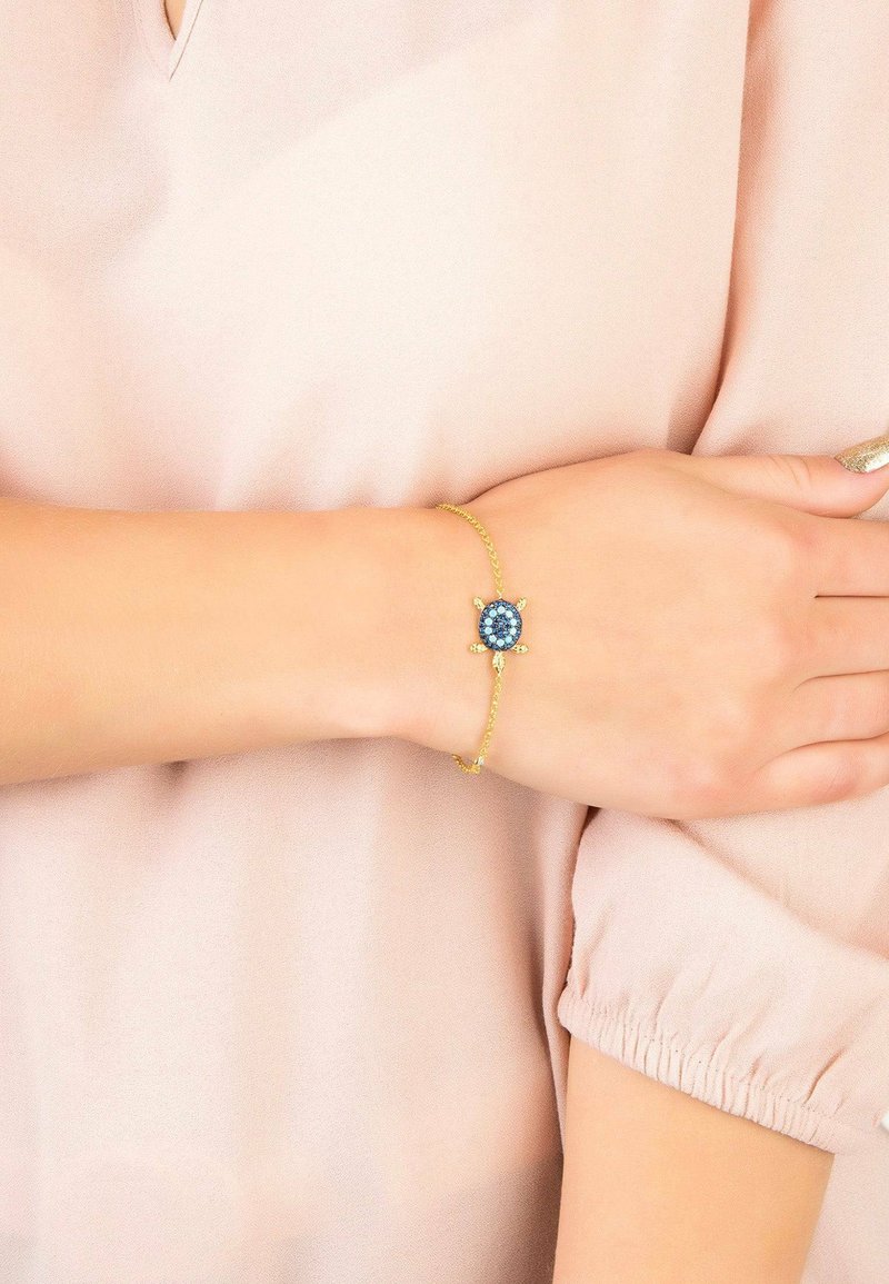 Bracciale in oro con una pietra blu centrale, circondata da piccoli dettagli blu, indossato su un polso scoperto su un morbido tessuto di colore chiaro.