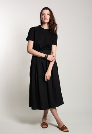 Zwarte katoen midi-jurk met korte mouwen, een aansluitende taille en een vloeiende rok. Gepaard met bruine platte sandalen.