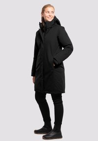 Abrigo largo negro con capucha, con cremallera frontal, bolsillos laterales y una tela suave y resistente al agua. Combinado con leggings negros y botas.