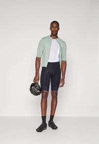 Kerékpáros öltözet világoszöld rövid ujjú mezből, fehér hálós aláöltözetből, fekete sortból és fekete kerékpáros cipőből, fekete sisakot tartva.