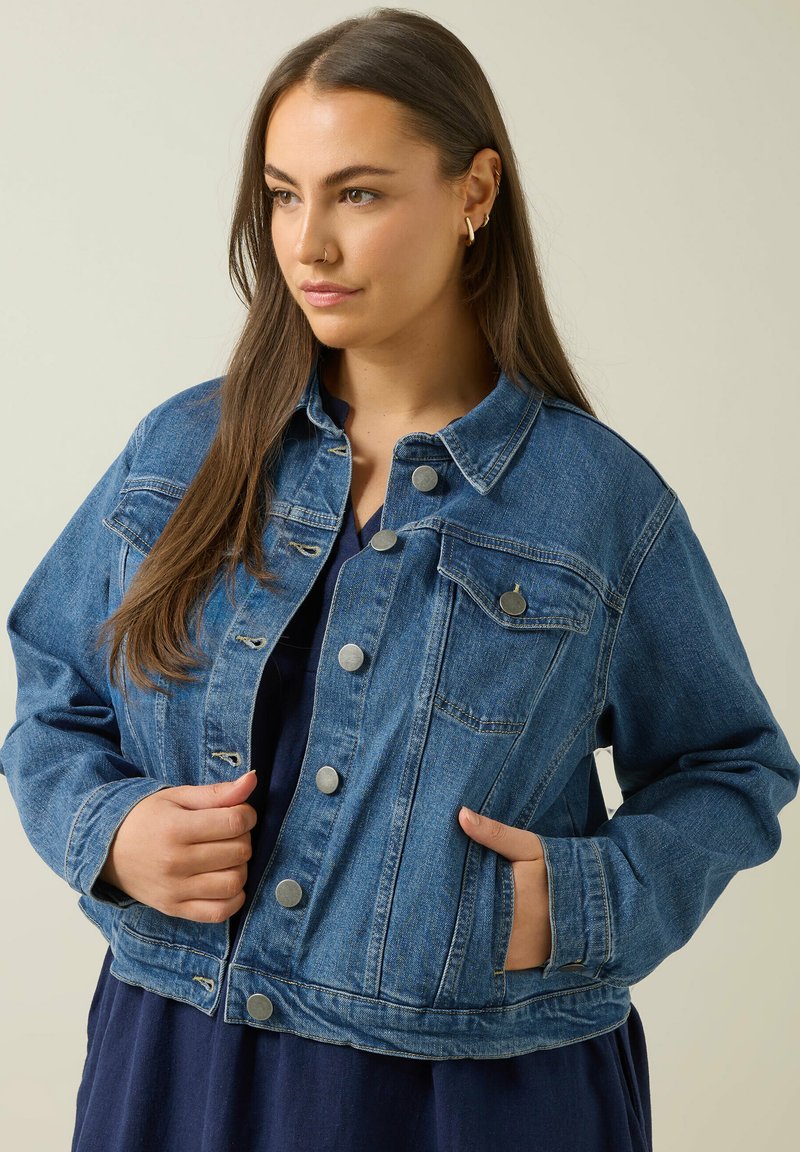 Jeansjacka i mellanblå, med knäppning framtill, två bröstfickor och en lös passform. Tyget har en slät textur och lätt blekning.