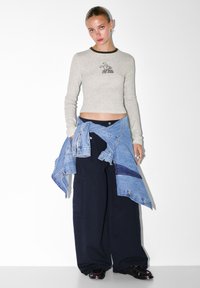 Svetlošedý top s dlhým rukávom a čiernym golierom a grafickou potlačou, skombinovaný s nadrozmernými námorníckymi nohavicami a viazanou denimovou bundou okolo pásu.