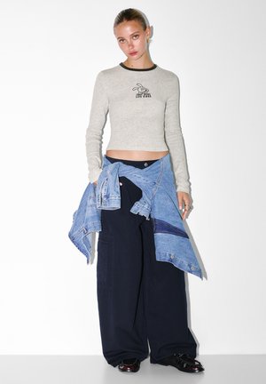 Hellgraues Langarmoberteil mit schwarzem Kragen und grafischem Druck, kombiniert mit übergroßen marineblauen Hosen und einer um die Taille gebundenen Jeansjacke.