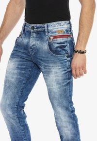 Blauwe denim jeans met een vervaagde wassing, voorzien van een rode ritszak, metalen hardware en een slim-fit ontwerp. Fluweelachtige textuur en klassieke snit.