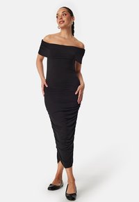 Robe noire à épaules dénudées avec une silhouette ajustée et une texture froncée, arrivant au milieu du mollet. Portée avec des ballerines noires, présentant une finition en tissu lisse.