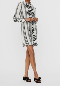 Svart och vit mönstrad skjortklänning med långa ärmar, krage och knytning i midjan; textil med struktur och geometriska mönster samt sidoklyftor.