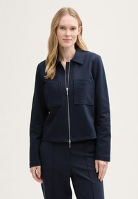 Mörkblå zip-up-jacka med två framfickor, tillverkad av texturerat tyg, med krage och långa ärmar, i kombination med matchande byxor.