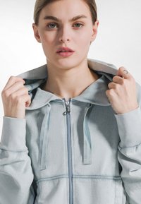 Lyseblå hættesweater med lynlås i blød tekstur, der har høj krave, snoretræk og kontrastlynlås med diskrete syningsdetaljer.