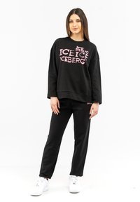 Schwarzer Sweatshirt mit pinkem "ICEBERG"-Text, lockere Passform und langen Ärmeln, kombiniert mit schwarzen Hosen und weißen Sneakers.