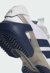 Chaussure de sport blanche avec des bandes bleu marine, une tige en mesh, une semelle en caoutchouc et un design texturé. Dispose d'un talon prononcé pour le maintien.