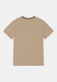 Camiseta de manga corta beige con el texto "Calvin Klein Jeans" en el cuello trasero, diseño liso, colocada sobre un fondo blanco.