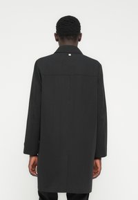 Μαύρο μακρύ παλτό από απαλό ύφασμα· διαθέτει γιακά, μακριά μανίκια και ένα μονό διακοσμητικό κουμπί στην πλάτη. Απλός και ευθύς σχεδιασμός.