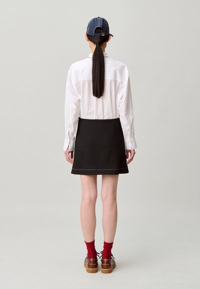 Gonna nera con dettaglio bordo a contrasto, camicia a righe bianche, e cappellino blu. Calzini rossi e scarpe marroni con lacci completano l'outfit.