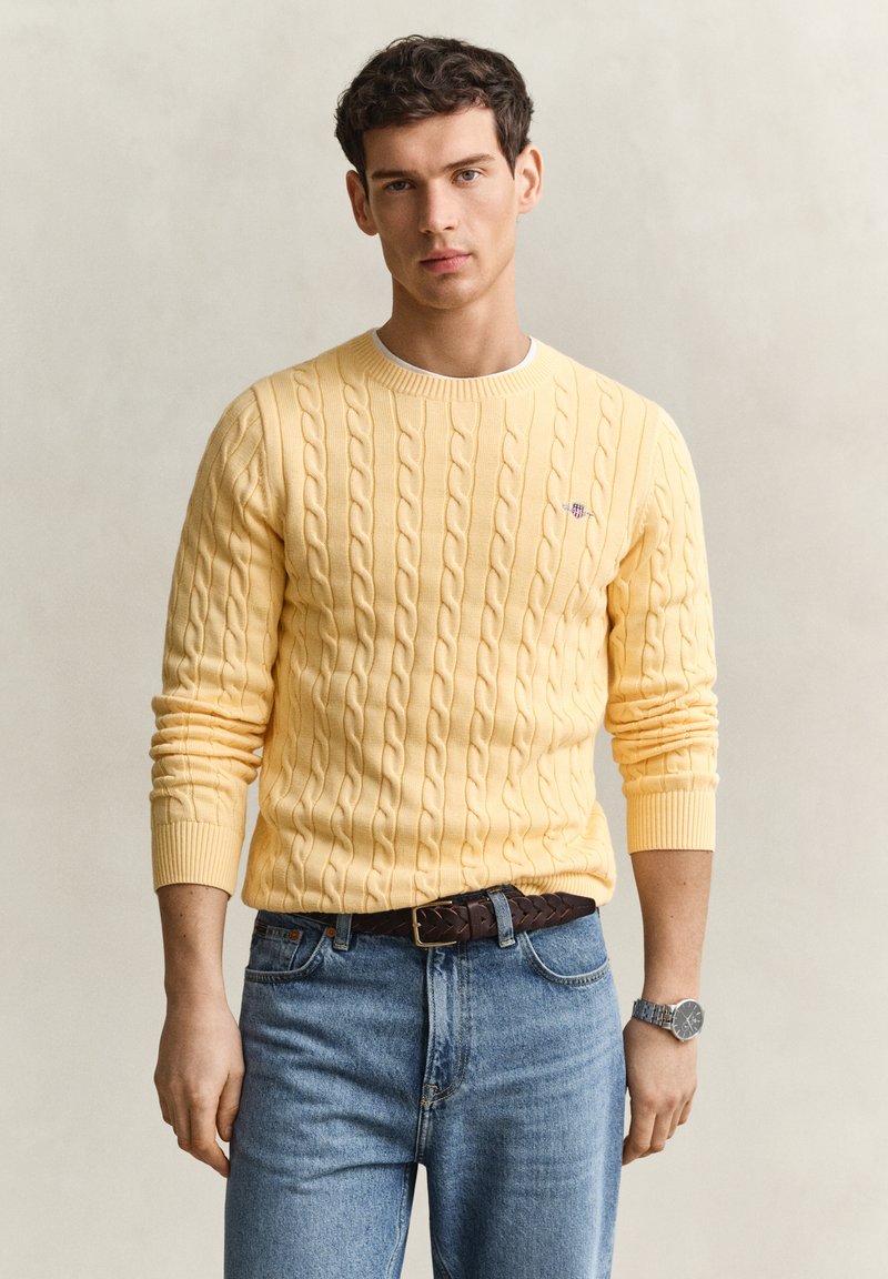 Homme portant un pull jaune torsadé, un jean bleu avec une ceinture tressée marron, et une montre-bracelet argentée, debout devant un fond neutre.